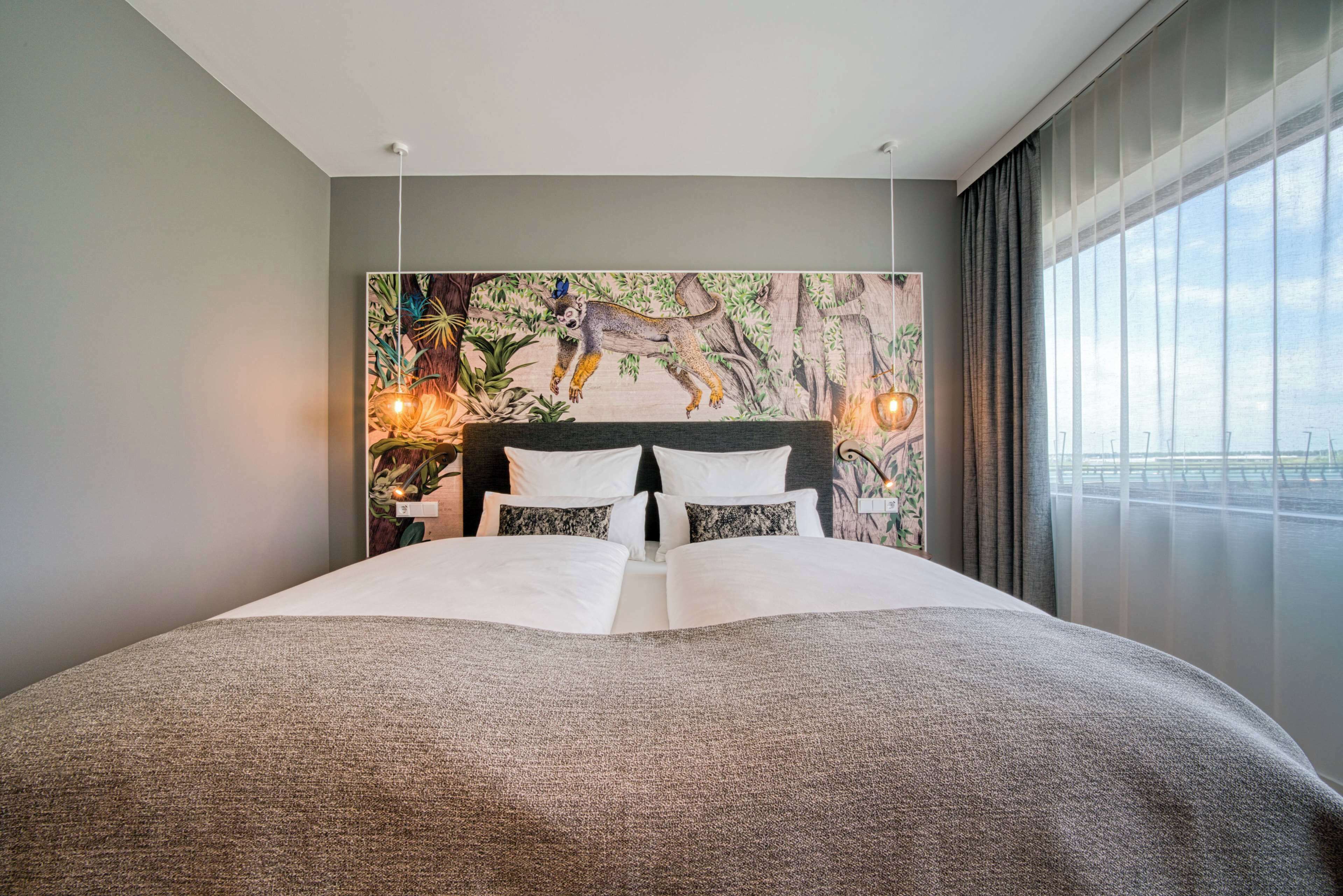 Una cama grande con ropa blanca y una manta gris, un mural de lémur en la pared y una ventana con cortinas.