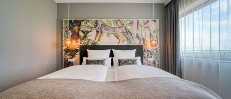 Una cama grande con ropa blanca y una manta gris, un mural de lémur en la pared y una ventana con cortinas.