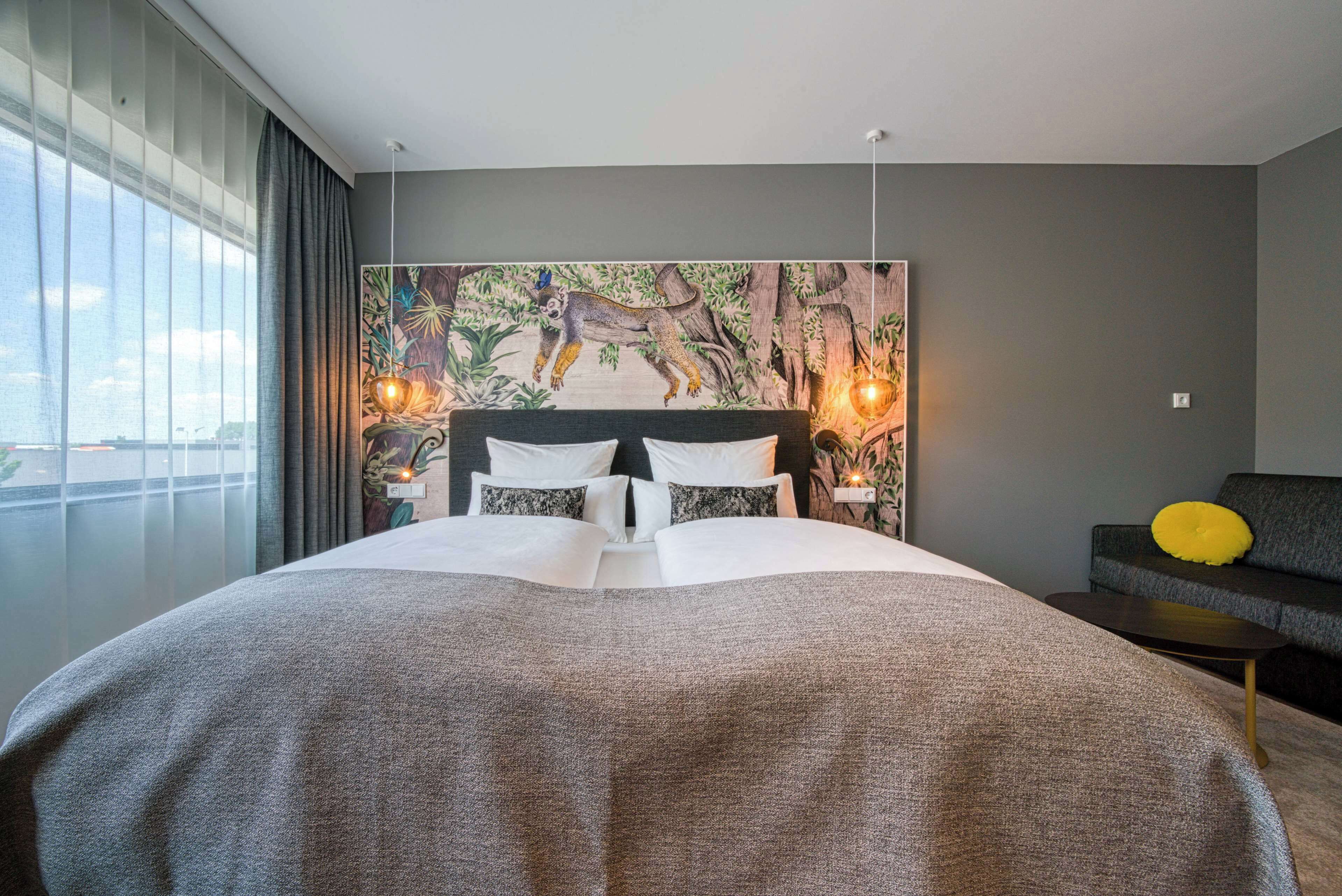Una habitación de hotel moderna con una cama grande, un cabecero con una obra de arte con motivos forestales, vistas a la ciudad y una mesita de noche con una lámpara.