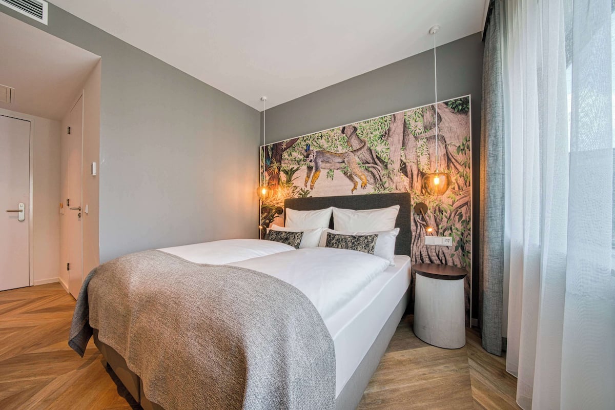 A neatly made bed with a grey blanket, a wall-mounted nightstand with a lamp, and a large nature-themed mural on the wall.
