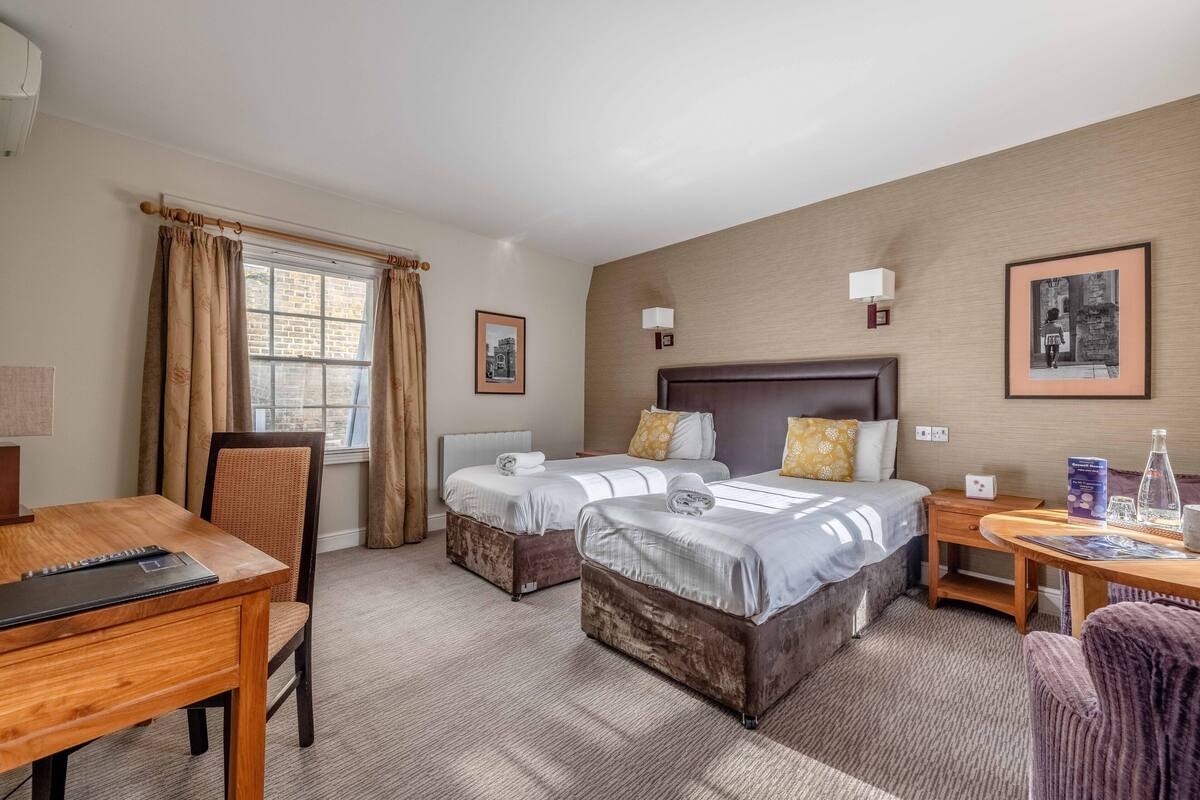 Quarto de hotel com duas camas, uma escrivaninha de madeira, uma cadeira, uma mesa pequena e uma janela com cortinas.