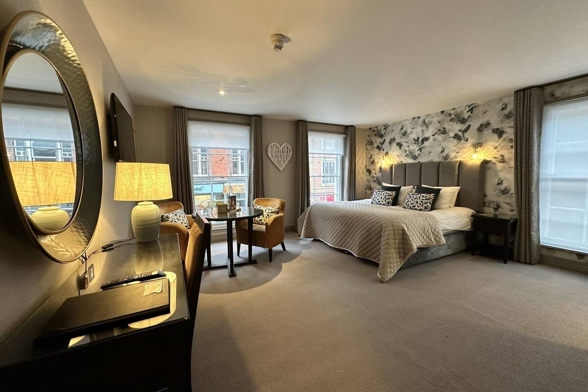 Quarto de hotel com uma cama grande, uma escrivaninha com luminária, uma cadeira e um espelho amplo.