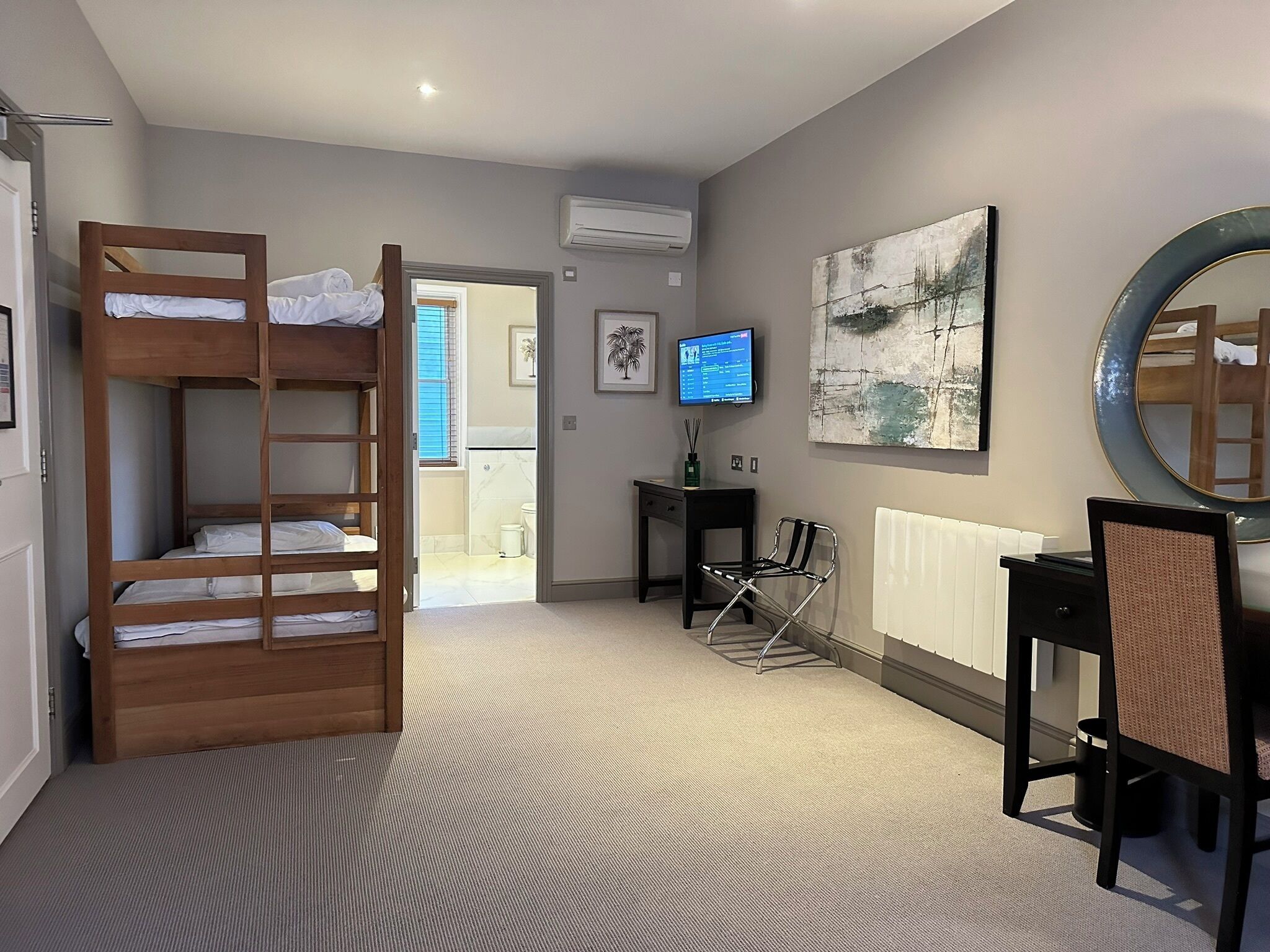 Quarto de hotel com beliche, escrivaninha com cadeira, televisão e um espelho grande.