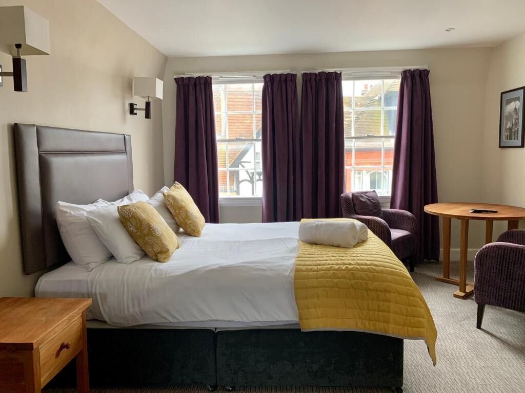 A hotel room with a bed, a bedside table, a chair, a small table, and a window with curtains.