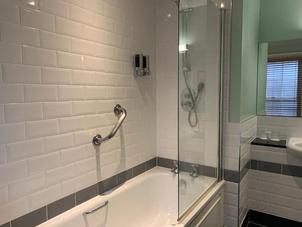 Une salle de bain moderne avec des carreaux de céramique blancs rectangulaires, une douche à parois de verre et une baignoire blanche équipée d’une barre d’appui.