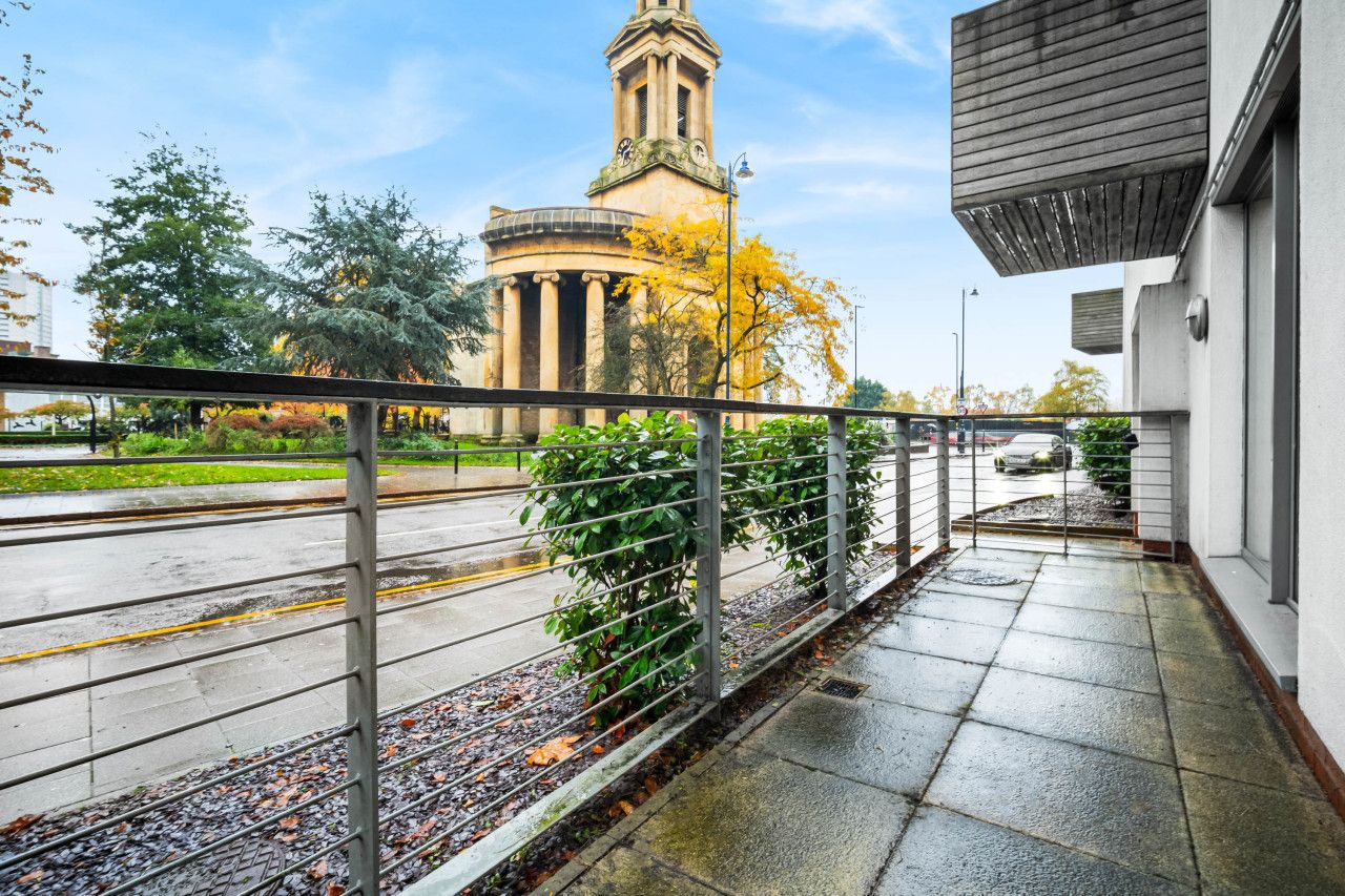A balcony with a metal railing overlooking a street with a historic building and trees in the background.