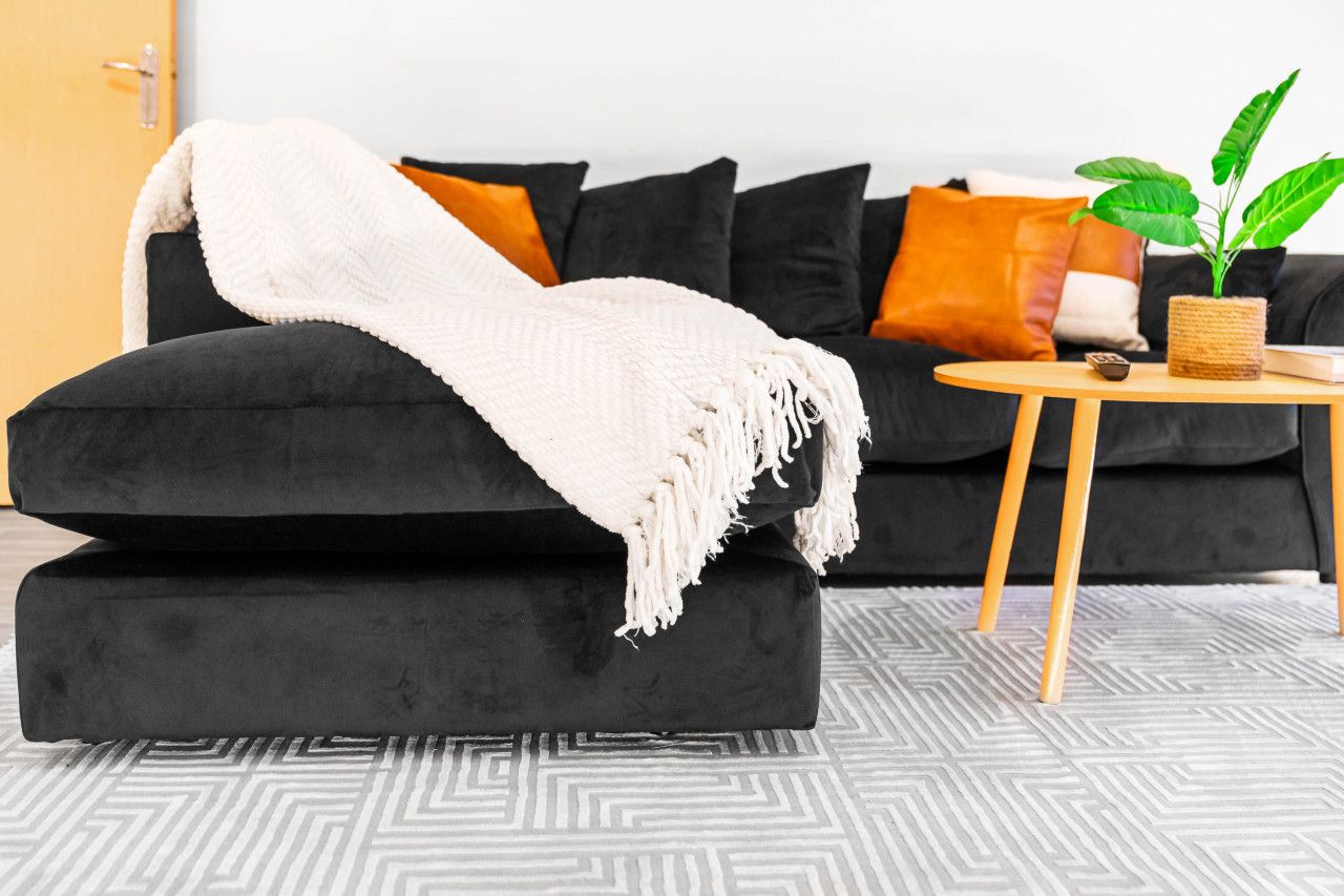 A black sofa with a white throw blanket, a small round wooden table with a plant, and a patterned rug.