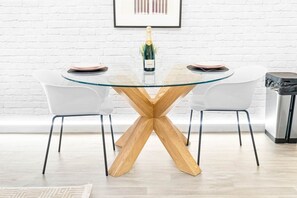 A round glass table with a wooden X-shaped base, two white chairs, a bottle of champagne, and a framed picture on the wall. - Stylish City Centre Apartment -  Terrace - Netflix (West Midlands)