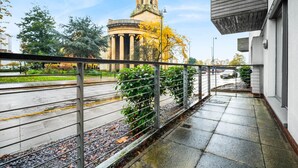 Un balcone con ringhiera in metallo che si affaccia su una strada con un edificio storico e alberi sullo sfondo.