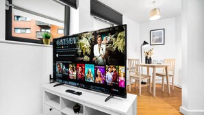 A modern living room with a flat-screen TV displaying the movie 'Gatsby'. There is a white entertainment unit below the TV, a dining area with a wooden table and chairs, and a window with a view of buildings outside. - Birmingham City Centre Apartment - SuperHost
 (Birmingham)