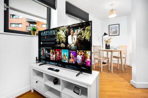 A modern living room with a flat-screen TV displaying the movie 'Gatsby'. There is a white entertainment unit below the TV, a dining area with a wooden table and chairs, and a window with a view of buildings outside.