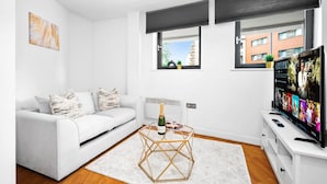 A modern living room with a white sofa, a glass coffee table, a flat-screen TV, and a large window with a view of buildings. - Birmingham City Centre Apartment - SuperHost
 (Birmingham)