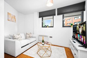 A modern living room with a white sofa, a glass coffee table, a flat-screen TV, and a large window with a view of buildings.