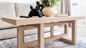 Ein Holztisch mit einer Vase und einem Buch darauf, platziert in einem Wohnzimmer mit einer Couch und einer Pflanze im Hintergrund.