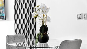 Ein moderner Essbereich mit einem Glastisch, zwei Stühlen, einer schwarzen Vase mit weißen Blumen und einer dekorativen Wandverkleidung.