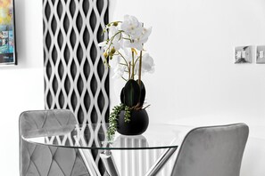 Ein moderner Essbereich mit einem Glastisch, zwei Stühlen, einer schwarzen Vase mit weißen Blumen und einer dekorativen Wandverkleidung.