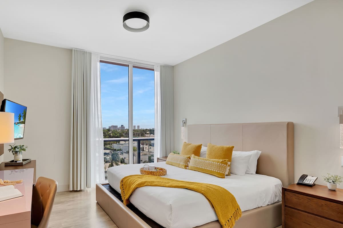 A modern hotel room with a large bed, a desk, a chair, a TV, and a view of the city through large windows.