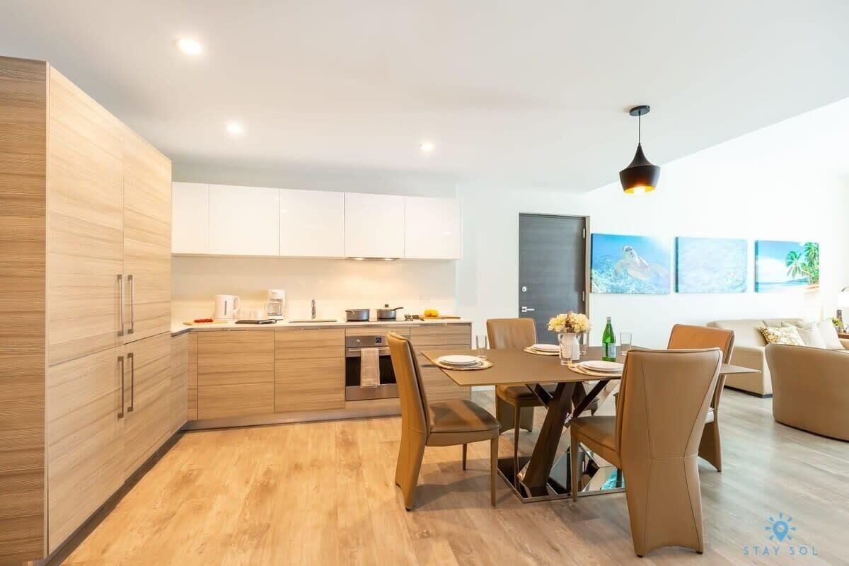 Una cocina moderna con armarios de madera, un área de comedor con mesa y sillas, y un salón con sofá y una obra de arte en la pared.