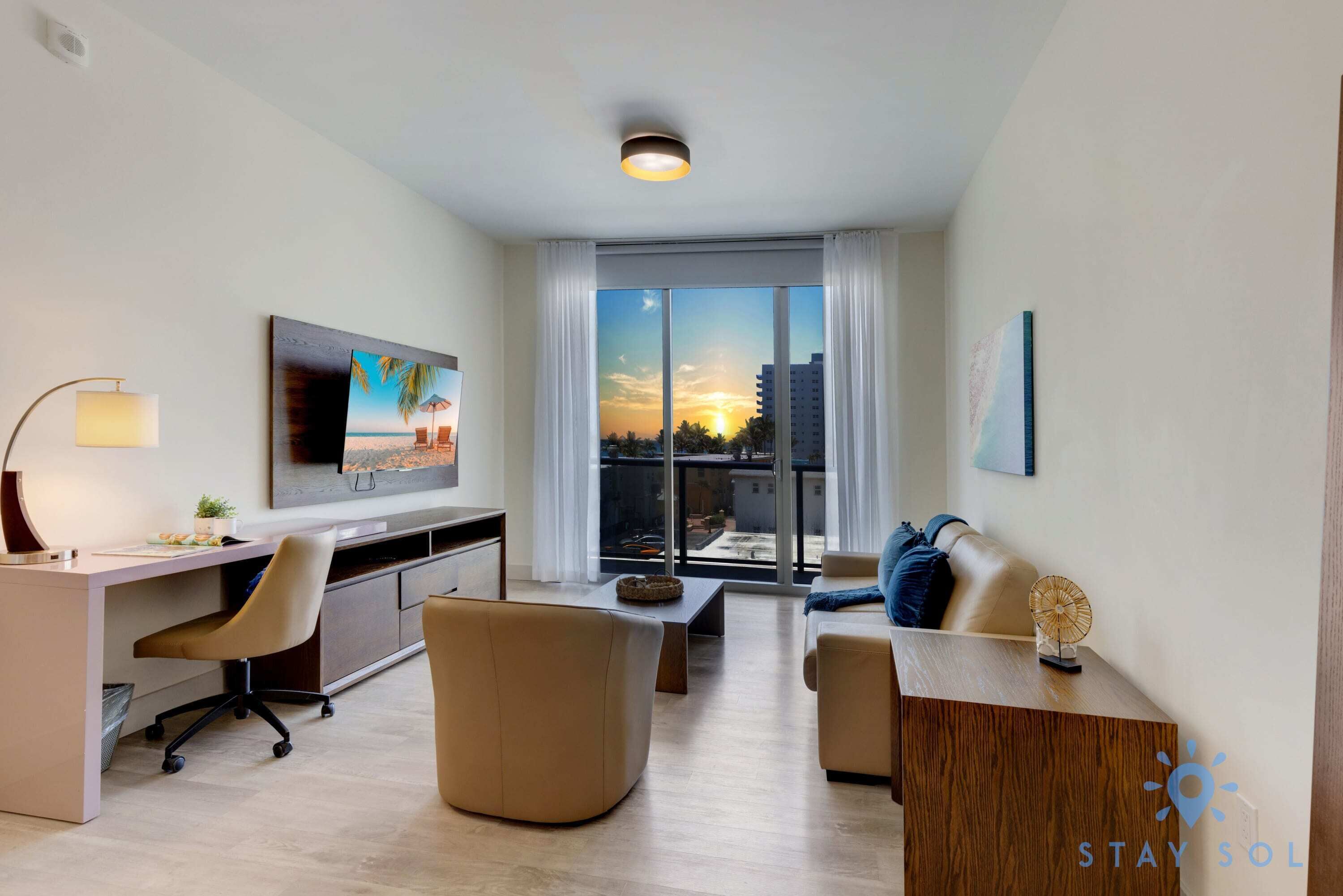 Uma sala de estar moderna com uma escrivaninha, assentos confortáveis, uma janela grande com vista para o pôr do sol e uma TV de tela plana na parede.