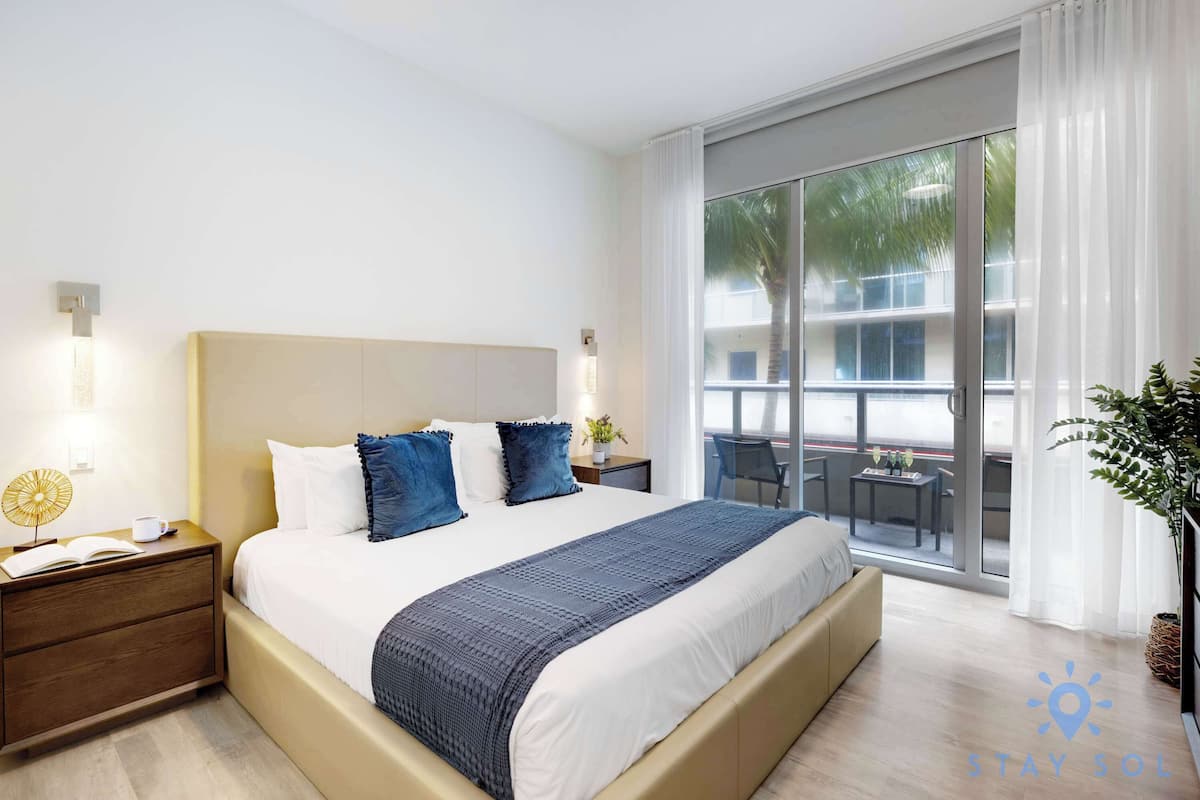 A modern hotel room with a large bed, a bedside table with a lamp, and a view of the outside balcony.