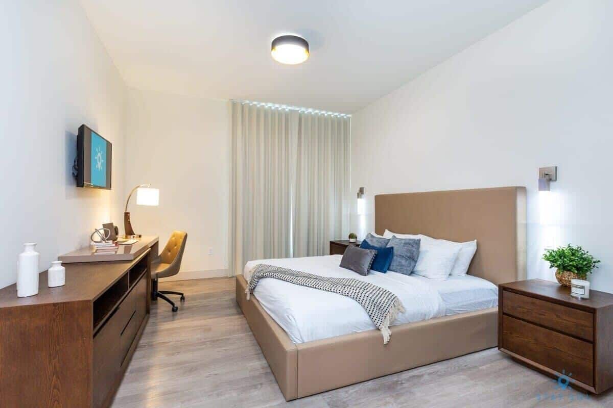 A modern hotel room with a bed, bedside tables, a desk, a chair, and a large window with curtains.