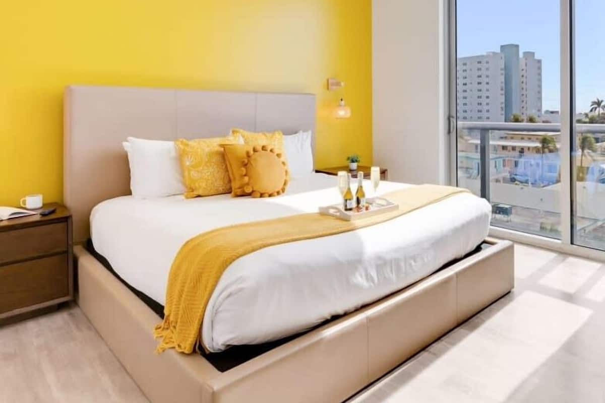 A hotel room with a large bed, a nightstand, a bedside table, a window with a view of buildings, and a yellow wall.