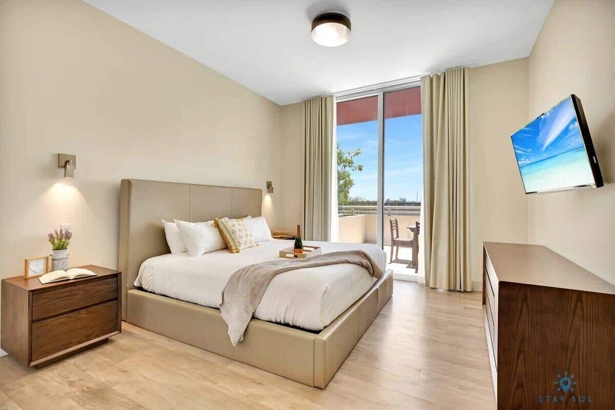 A modern hotel room with a large bed, a desk, a flat-screen TV, and a balcony with curtains.