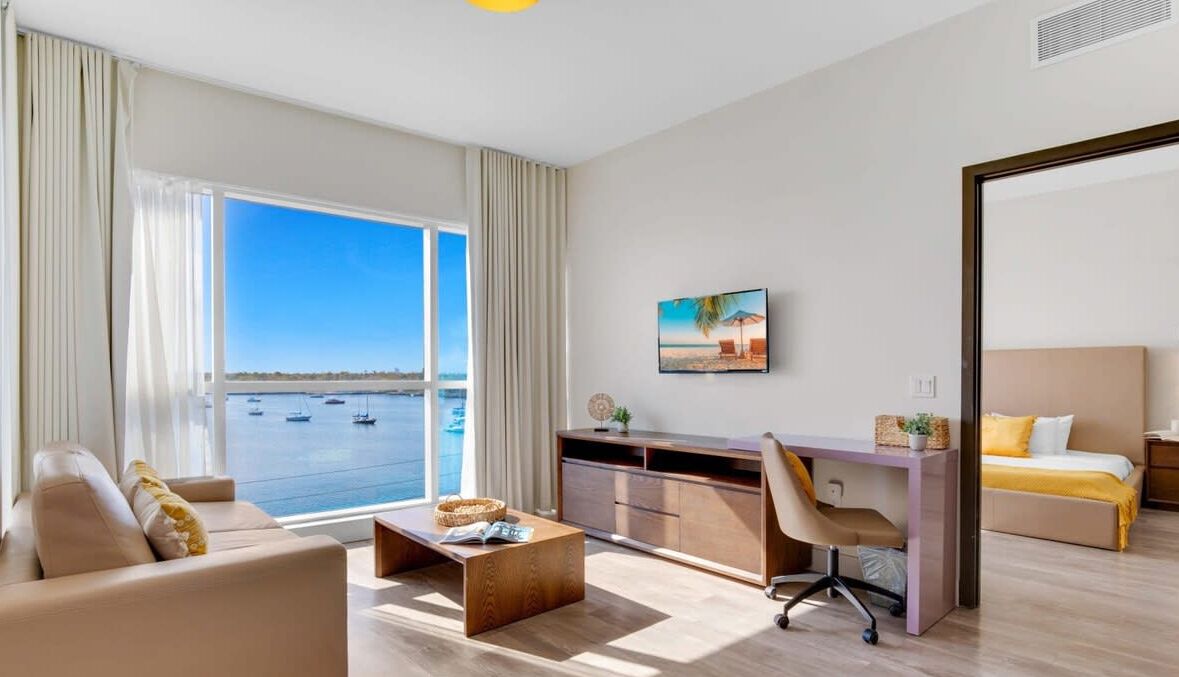 Ett modernt hotellrum med ett stort fönster som vetter mot havet, en bekväm soffa, ett skrivbord med stol och en säng med en gul detaljfärg.