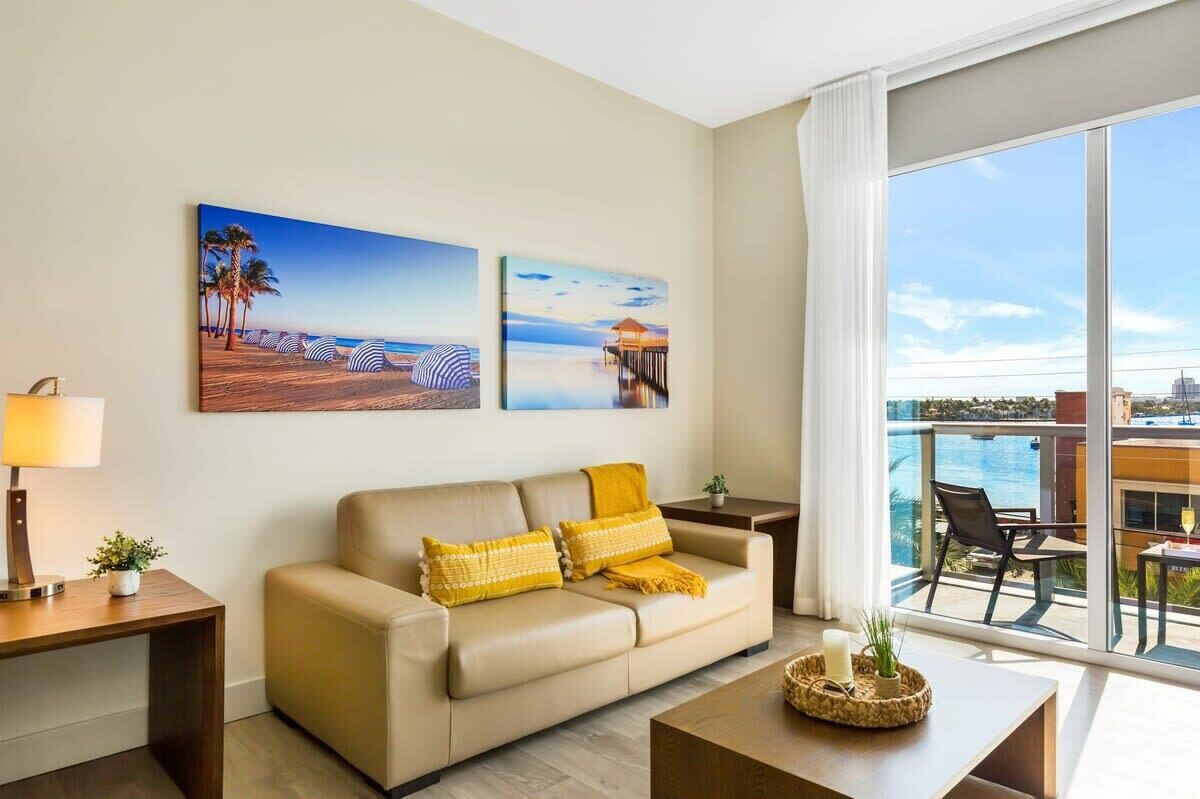 Une chambre d’hôtel moderne avec un canapé beige, deux tableaux représentant des scènes de plage, un balcon avec vue sur la mer et une petite table avec une bougie.