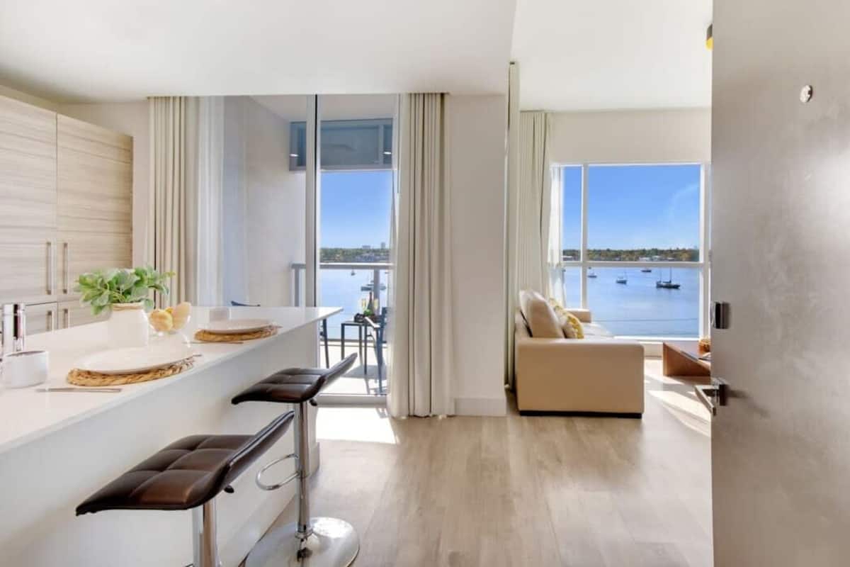 A modern kitchen with a large island, a dining area with bar stools, a sofa by a large window overlooking a body of water, and a view of boats.
