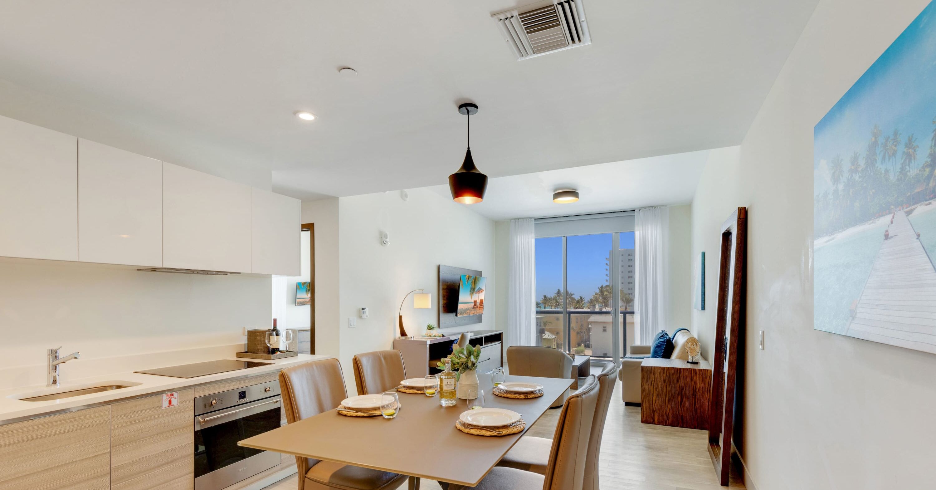 A modern kitchen with white cabinets, a dining area with a wooden table, and a large window offering a view of palm trees.