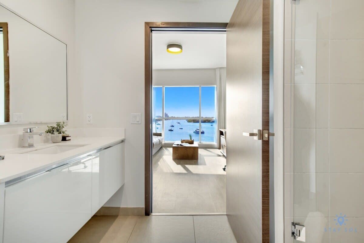 A modern bathroom with a large mirror, white tiles, and a view of the sea through a large window.