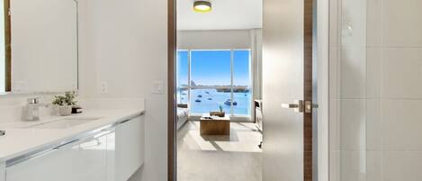 A modern bathroom with a large mirror, white tiles, and a view of the sea through a large window.
