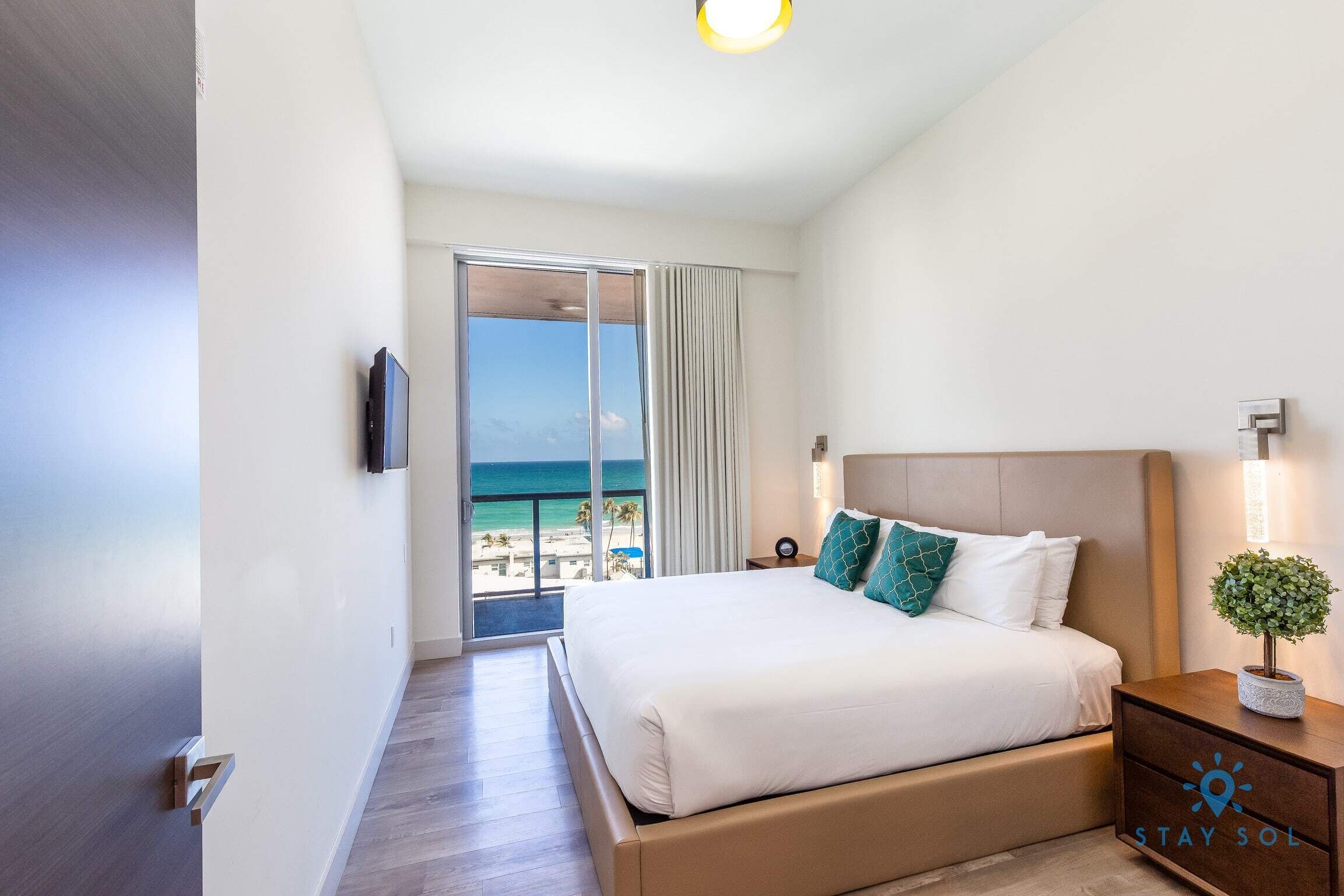 A hotel room with a bed, a nightstand, a TV, a window with a view of the beach, and a balcony.