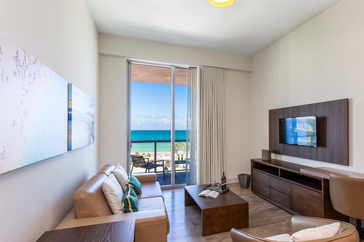A modern living room with a sofa, coffee table, TV, and a balcony with a view of the beach.