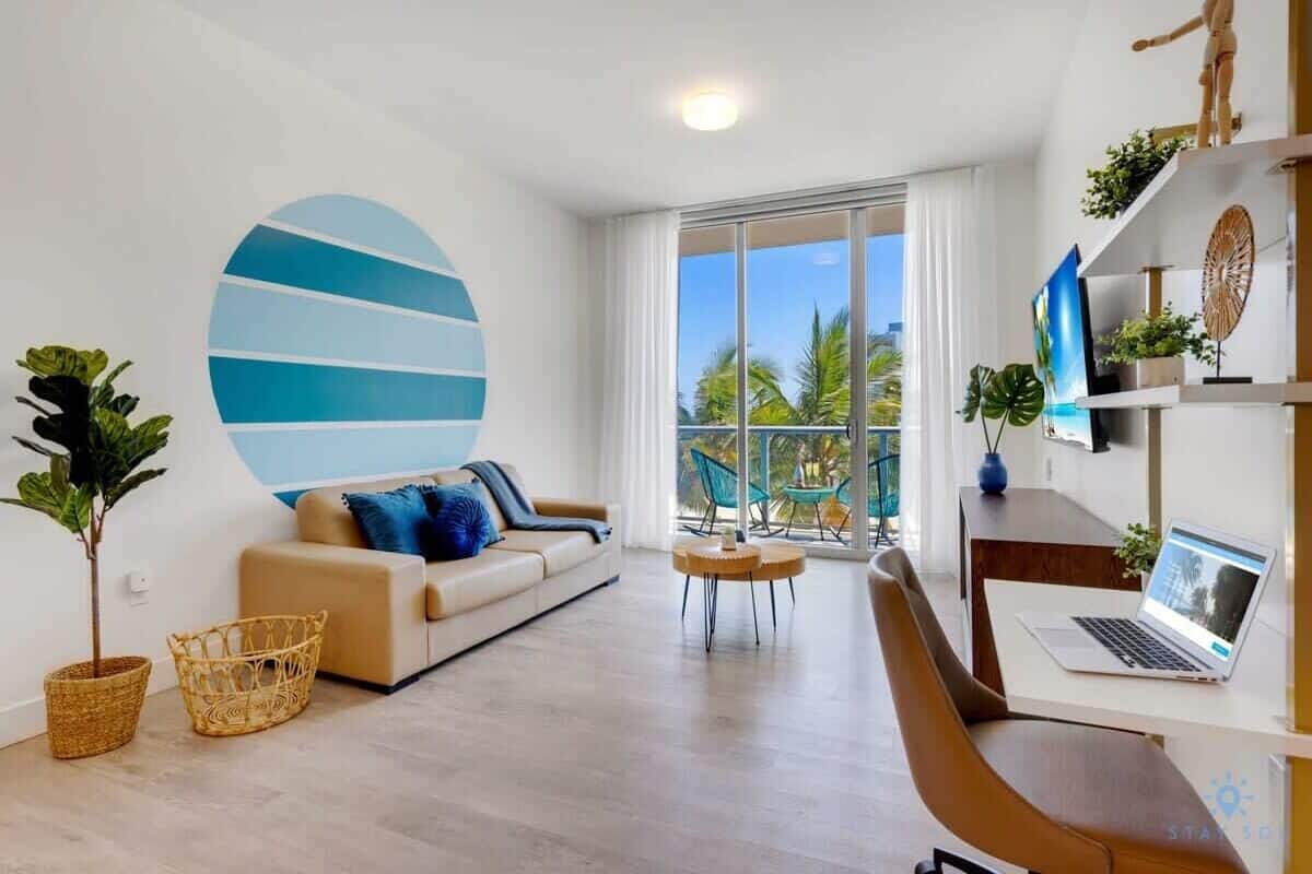 A modern living room with a sofa, a round coffee table, a laptop on a desk, and a large window with a view of palm trees.