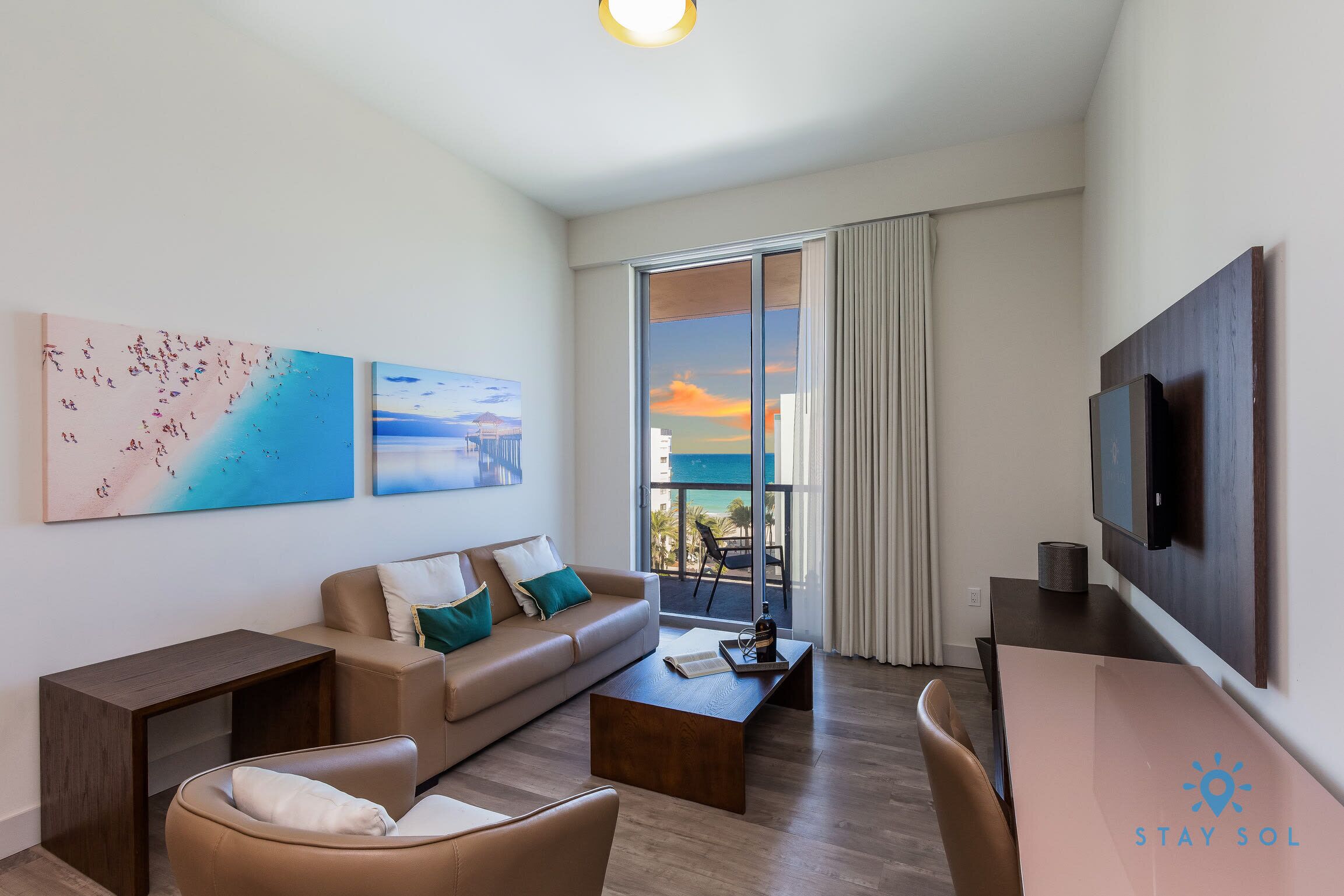 Un salon moderne avec un canapé, une table basse et un téléviseur. Les murs sont ornés de tableaux et un balcon offre une vue agréable.