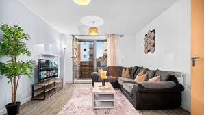 Ein modernes Wohnzimmer mit einer Couch, einem Couchtisch und einem Fernseher. Eine Pflanze, ein Teppich und eine Tür zu einem Balkon sind ebenfalls vorhanden.