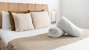 Un letto rifatto con cura, con una testiera beige, cuscini bianchi e marroni e un asciugamano bianco arrotolato su una coperta beige.