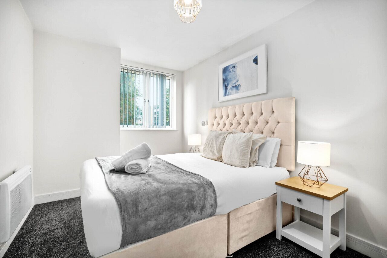 Een moderne slaapkamer met een gebloemd hoofdeinde, een nachtkastje met een lamp, en een raam met jaloezieën.
