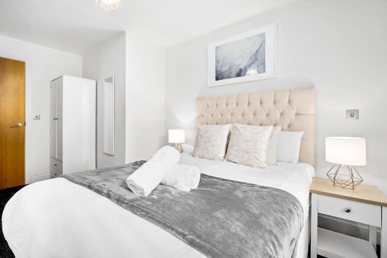 Quarto de hotel com uma cama grande, duas mesas de cabeceira com abajures, uma porta e uma obra de arte emoldurada na parede.