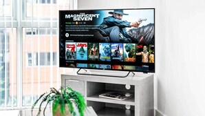 Et fladskærms-tv på et moderne stativ viser en film, med en plante og et magasin på et bord foran.