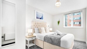 Et soveværelse med en betrukken sengegavl, et stort vindue med skodder, et sengebord med en lampe og et billede i ramme på væggen.