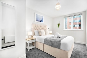 Ein Schlafzimmer mit einer gepolsterten Kopfteile, einem großen Fenster mit Jalousien, einem Nachttisch mit Lampe und einem gerahmten Bild an der Wand.