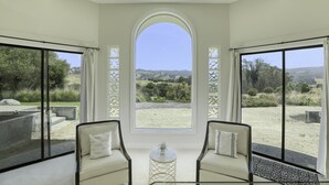 A room with a large arched window, two chairs, a small table, and a view of a landscaped garden. - Avila Beach Estate: Exceptional Hope Ranch (San Luis Obispo)