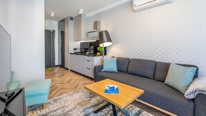 Een moderne woonkamer met een bank, een salontafel en een televisie. In de achtergrond is een keuken met een fornuis en kastjes te zien.
