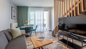 Een moderne woonkamer met een bank, salontafel en televisie. Er is een zithoek met stoelen en een tafel. Een trap met houten leuningen is zichtbaar.