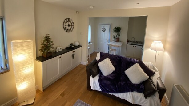 A cozy living room with a sofa, a lamp, a clock, and a kitchen area in the background. - Converted Stables - Pet Friendly - Enclosed Garden (Pontefract)