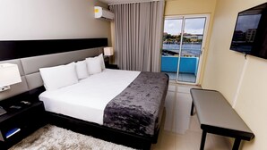 Une chambre d’hôtel avec un grand lit, une table de chevet avec une lampe, un téléviseur fixé au mur et une vue sur un plan d’eau, accessible par une porte coulissante.