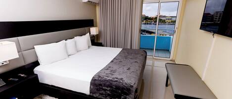 Habitación de hotel con una cama grande, una mesita de noche con lámpara, televisor montado en la pared y vista a una masa de agua a través de una puerta corrediza.